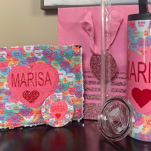 May include: A pink and red Valentine's Day gift set with the name "MARISA" on a tumbler, a sequin pillow, and a round coaster. The items are decorated with candy hearts with various messages.