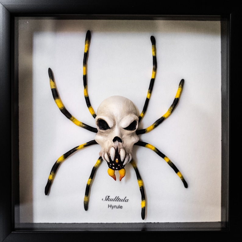 May include: A framed black and yellow spider with a skull-like head. The spider is labeled "Skulltula Hyrule".