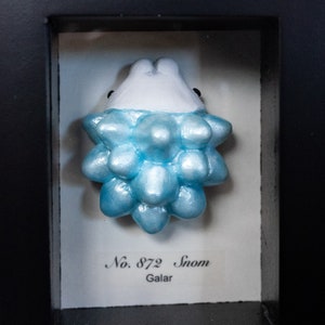 Może przedstawiać: Niebieska i biała figurka Pokemona o imieniu Snom. Figurka znajduje się w gablocie z białą etykietą, na której widnieje napis "No. 872 Snom Galar".