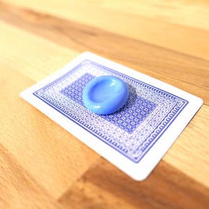 Puede incluir: Un objeto azul claro, de forma ovalada, descansa sobre una carta de juego. La carta tiene un diseño azul y blanco. El fondo es una superficie de madera.