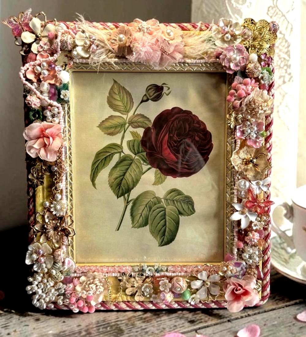 May include: Ornate picture frame with a botanical print of a dark red rose. The frame is embellished with pearls, fabric flowers, and gold accents. The frame is rectangular and has a vintage aesthetic.