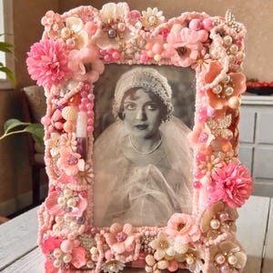 May include: A pink, ornate picture frame with a black and white photo of a woman. The frame is heavily decorated with pink flowers, pearls, beads, and rhinestones. A small tube of lip gloss is attached to the side of the frame.