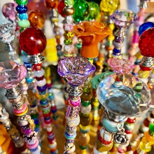May include: A collection of colorful beaded wands with various decorative tops, including flowers, crystals, and beads. The wands are made with a variety of materials, including glass, metal, and plastic.