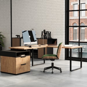 Könnte beinhalten: Ein moderner Büroarbeitsplatz mit einem großen L-förmigen Schreibtisch aus Holz mit schwarzen Metallbeinen. Der Schreibtisch verfügt über einen Computermonitor, einen Laptop und einen grünen Bürostuhl. Der Raum hat eine weiße Backsteinwand und große Fenster.