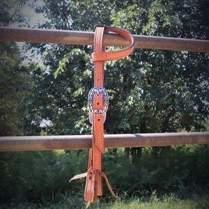 May include: A tan leather horse bridle with a decorative silver buckle. The bridle is hanging on a brown metal fence with a green background of trees and grass. The bridle is made of stitched leather.