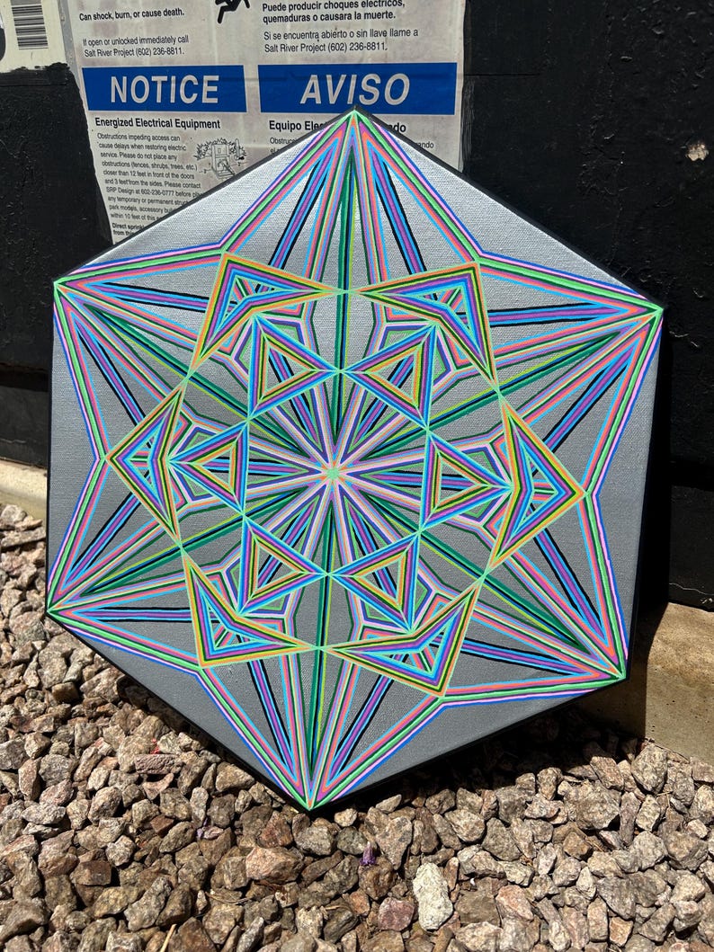 May include: A hexagonal artwork with a silver surface, featuring a complex geometric design. The artwork displays a central starburst pattern with radiating lines in blue, green, pink, and yellow. The artwork is placed on a bed of small rocks. The artwork is placed on a bed of small rocks.