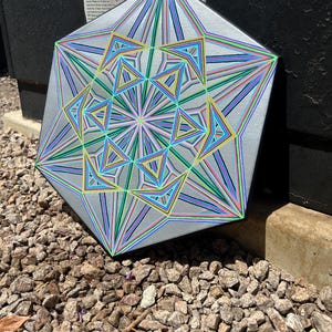 May include: A geometric artwork with a silver background and colourful lines in shades of blue, green, yellow, and pink. The artwork is a multi-sided shape with a complex, layered design. The artwork is resting on a bed of small rocks.
