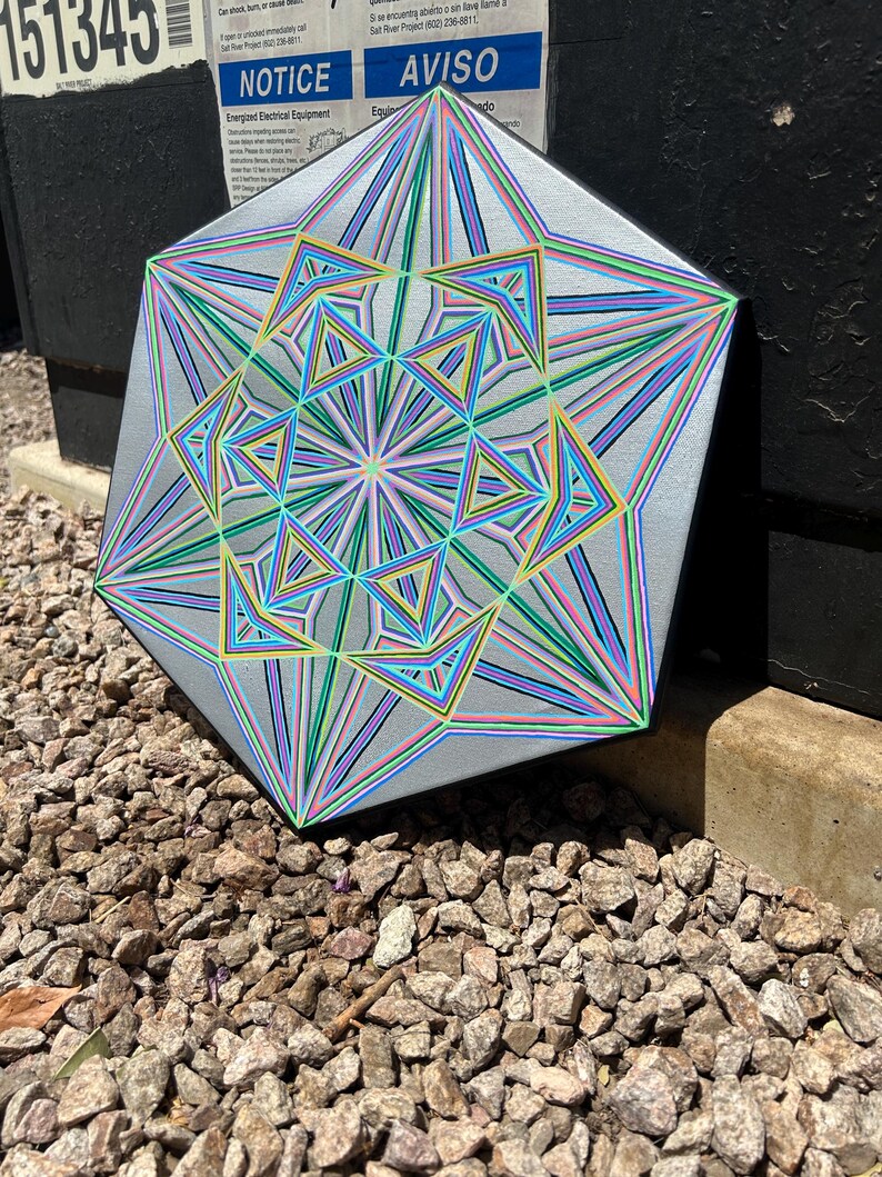 May include: A geometric artwork with a silver background and colourful lines in shades of blue, green, yellow, and pink. The artwork is a multi-sided shape with a complex, layered design. The artwork is resting on a bed of small rocks.