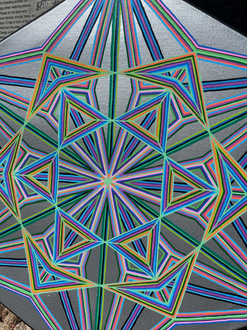May include: Abstract geometric artwork on a silver background. The design features a symmetrical pattern of colourful lines in shades of blue, green, pink, and orange, creating a starburst effect. The artwork is on a square canvas.