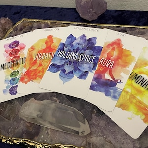 May include: A collection of oracle cards with watercolor illustrations and text. The cards feature words like "Meditation," "Vibration," and "Holding Space." The color palette includes blues, oranges, and yellows. A clear quartz crystal and amethyst are also present.