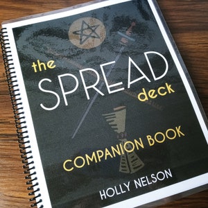 以下が含まれることがあります： 「The Spread Deck」タロットデッキのコンパニオンブックの表紙。表紙は黒で、黄色のテキストと五芒星が描かれている。テキストには「The Spread Deck」と「Companion Book」と、著者の名前「Holly Nelson」が書かれている。