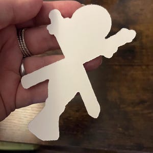 以下が含まれることがあります： 両手を広げ、片足を前に出した人の形をした白い紙の切り抜き。切り抜きは、指に複数のリングが付いた手で持たれています。背景は濃い茶色の木製の表面です。