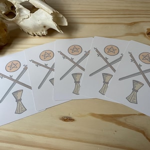 May include: A collection of tarot cards featuring illustrations of crossed swords, a staff, and a chalice. Each card has a circular emblem with a star. The cards are arranged on a wooden surface, with a skull and crystal in the background.