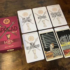 May include: A tarot card deck titled "the SPREAD deck LOVE" with a burgundy box and several tarot cards. The cards have text such as "ARE THEY CHEATING?" and "RED FLAGS". The deck is by Holly Nelson.