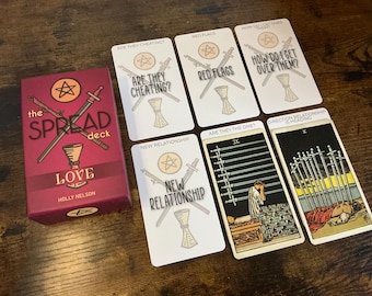 Puede incluir: Una baraja de cartas de tarot titulada "the SPREAD deck LOVE" con una caja burdeos y varias cartas de tarot. Las cartas tienen texto como "ARE THEY CHEATING?" y "RED FLAGS". La baraja es de Holly Nelson.