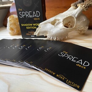May include: A deck of black cards with gold lettering that reads "The Spread Deck Shadow Work Edition". The cards are fanned out on a wooden surface. A white animal skull with antlers is in the background.