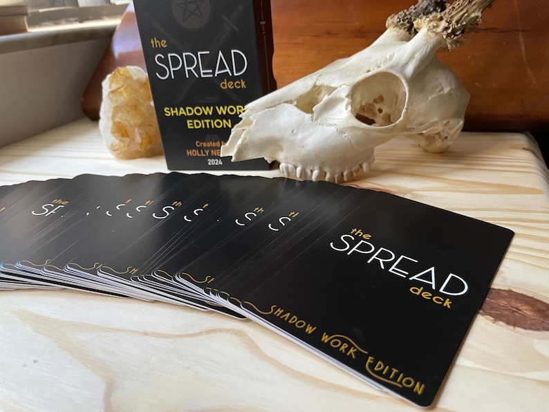 May include: A deck of black cards with gold lettering that reads "The Spread Deck Shadow Work Edition". The cards are fanned out on a wooden surface. A white animal skull with antlers is in the background.