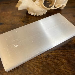 以下が含まれることがあります： 長方形で半透明のセレナイト充電プレート。プレートは明るい真珠のような白色で、滑らかで研磨された表面を持っています。背景には頭蓋骨が見えます。このアイテムは、クリスタルの浄化やエネルギーワークによく使用されます。