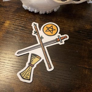 以下が含まれることがあります： タロットカードのシンボルが描かれたステッカー。剣、杖、聖杯、星の入った金の円が描かれています。ステッカーは白い縁取りで、濃い木目の背景に配置されています。