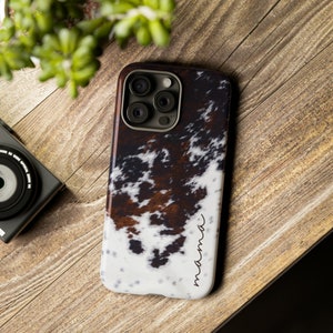 May include: A white, brown and black cowhide patterned phone case with a black camera cutout and the word "mama" written in cursive on the bottom right corner.