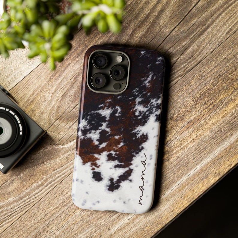 May include: A white, brown and black cowhide patterned phone case with a black camera cutout and the word "mama" written in cursive on the bottom right corner.