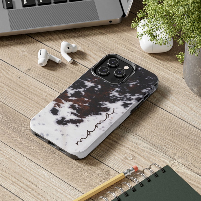 May include: A white phone case with a brown and black cowhide pattern. The case has a black camera bump and the word "mama" written in cursive on the bottom.