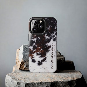 May include: A black phone case with a white and brown cowhide pattern. The case has a camera lens cutout and the word "mama" written on the side.