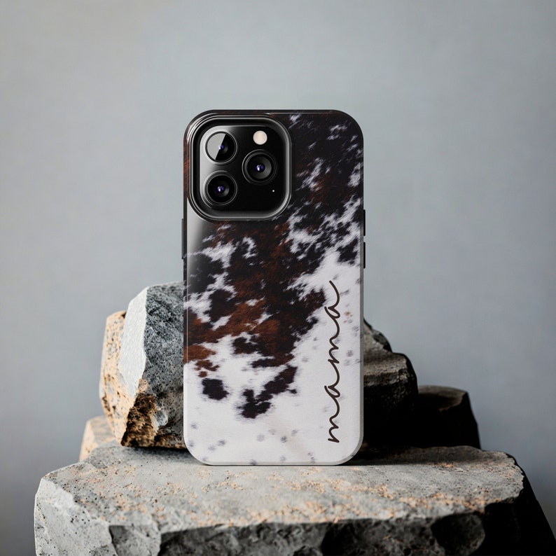 May include: A black phone case with a white and brown cowhide pattern. The case has a camera lens cutout and the word "mama" written on the side.