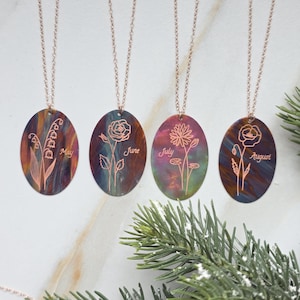 Może przedstawiać: Cztery owalne naszyjniki z kwiatowymi wzorami i wygrawerowanymi miesiącami maj, czerwiec, lipiec i sierpień. Wisiorki są przymocowane do delikatnych łańcuszków w kolorze różowego złota. Tło ma efekt marmuru z odcieniami niebieskiego, fioletowego i złotego.
