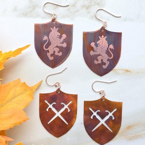 May include: Two pairs of shield-shaped copper earrings with a dark patina. One pair features an etched design of a crowned lion, while the other pair displays a cutout pattern of two crossed swords. Each earring hangs from a simple metal hook.