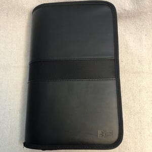May include: A black, rectangular case with a textured, woven band across the center. The case has a smooth, leather-like appearance and a black trim. The lower right corner features a small, embossed logo.