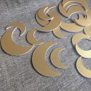 Puede incluir: Una colección de formas de media luna doradas con bordes blancos, dispersas sobre una superficie gris texturizada. Las lunas varían en tamaño, creando un tema celestial. Ideal para manualidades o proyectos decorativos.