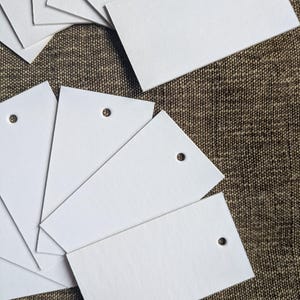 Puede incluir: Un conjunto de etiquetas rectangulares blancas en blanco con un pequeño agujero en la parte superior para ensartar. Las etiquetas están dispuestas sobre una superficie marrón texturizada, algunas se superponen. Las etiquetas son de papel grueso y mate.