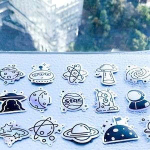 May include: A collection of 18 black and white space-themed stickers on a light blue background. The stickers feature various space-related designs, including planets, stars, rockets, aliens, and UFOs. The text "Interplanetary Astronauts" and "Universe" are featured on two of the stickers.