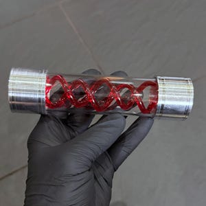 May include: A clear glass vial containing a red, double-helix structure. The vial is capped with silver-coloured metal at each end. The object is held by a gloved hand against a neutral background.