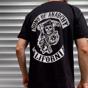 May include: Black t-shirt with a white graphic on the back. The graphic features a skull with a scythe, shield, and the words "SONS OF ANARCHY" and "CALIFORNI". The shirt is made of a soft material.