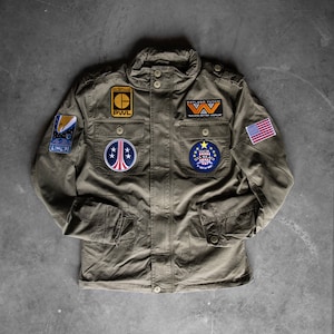 Op de afbeelding: Olijfgroene jas met meerdere patches, waaronder een Amerikaanse vlag, een USCM-patch en een Weyland-Yutani-patch. De jas heeft een knoopsluiting, twee borstzakken en twee onderste zakken. Militaire stijl.
