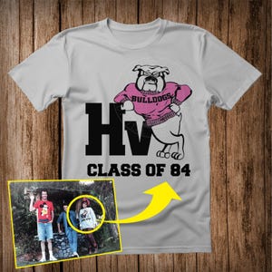 May include: A pale grey t-shirt with a bulldog mascot wearing a pink sweater, the word "Bulldogs" is visible. The shirt also displays "HV CLASS OF 84" in black block letters. A small photo is attached to the shirt.