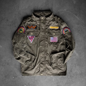 Op de afbeelding: Olijfgroene jas met meerdere patches, waaronder een Amerikaanse vlag, een regenboog en de tekst "WAYLAND YUTANI". De jas heeft een knoopsluiting, meerdere zakken en een hoge kraag. De jas is een militaire stijl.