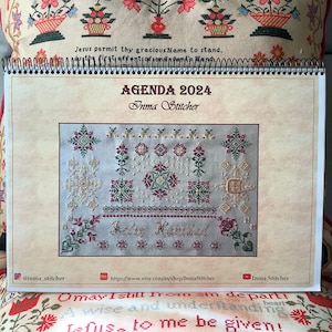 Puede incluir: Una agenda para 2024 con una cubierta beige que presenta un diseño de punto de cruz de una escena navideña con el texto "Feliz Navidad". La agenda se titula "AGENDA 2024" con el nombre "Inma Stitcher" debajo del título.
