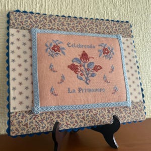Puede incluir: Una bordado de punto de cruz enmarcado con un fondo rosa y diseños florales azules y rojos. El texto "Celebrando La Primavera" está bordado en hilo azul.