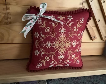 Op de afbeelding: Een rood kussen met een wit en gouden geborduurd ontwerp. Het kussen heeft een wit lint met groene en rode accenten eromheen gebonden.