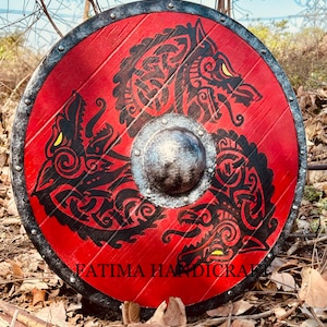 K&ouml;nnte beinhalten: Ein runder Holzschild mit einem schwarzen Metallrand und rotem Hintergrund. Der Schild zeigt ein schwarzes und gelbes Design mit drei Wolfsk&ouml;pfen mit komplizierten Mustern. Der Text "FATIMA HANDICRAFT" ist auf der Unterseite des Schildes aufgedruckt.