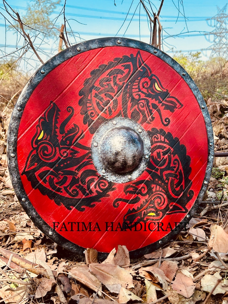 K&ouml;nnte beinhalten: Ein runder Holzschild mit einem schwarzen Metallrand und rotem Hintergrund. Der Schild zeigt ein schwarzes und gelbes Design mit drei Wolfsk&ouml;pfen mit komplizierten Mustern. Der Text "FATIMA HANDICRAFT" ist auf der Unterseite des Schildes aufgedruckt.