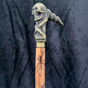 Może przedstawiać: Dekoracyjna laska do chodzenia z uchwytem w kształcie czaszki i skrzyżowanych kości w kolorze brązu. Drewniany trzon ma wzór gałęzi drzew i złoty pasek. Stylowy dodatek.