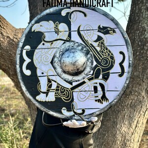 K&ouml;nnte beinhalten: Ein runder Holzschild mit einem schwarz-wei&szlig;en Design. Der Schild zeigt auf jeder Seite einen stilisierten Drachen und einen Vogel, die durch einen zentralen Metallboss getrennt sind. Der Schild ist mit goldenen Akzenten bemalt und hat einen Lederriemen.