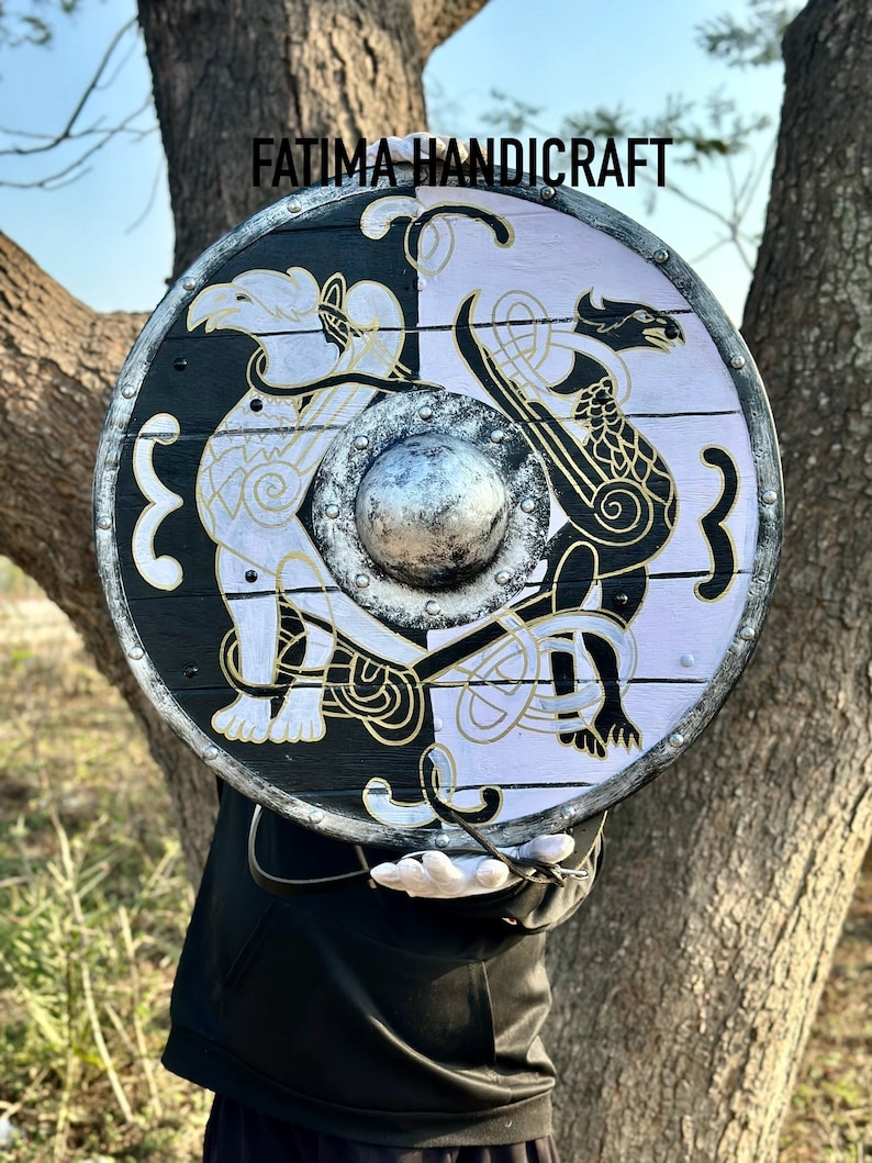 K&ouml;nnte beinhalten: Ein runder Holzschild mit einem schwarz-wei&szlig;en Design. Der Schild zeigt auf jeder Seite einen stilisierten Drachen und einen Vogel, die durch einen zentralen Metallboss getrennt sind. Der Schild ist mit goldenen Akzenten bemalt und hat einen Lederriemen.