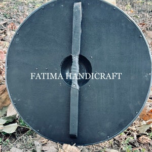 K&ouml;nnte beinhalten: Ein schwarzer Holzschild mit einem Ledergriff. Der Schild ist rund und hat einen Durchmesser von etwa 61 cm. Der Schild ist mit Metallnieten am Rand verziert. Der Text "FATIMA HANDICRAFT" ist auf den Schild gedruckt.