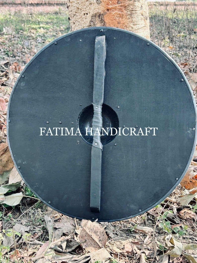 K&ouml;nnte beinhalten: Ein schwarzer Holzschild mit einem Ledergriff. Der Schild ist rund und hat einen Durchmesser von etwa 61 cm. Der Schild ist mit Metallnieten am Rand verziert. Der Text "FATIMA HANDICRAFT" ist auf den Schild gedruckt.