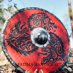 K&ouml;nnte beinhalten: Ein roter Holzschild mit einem schwarz-wei&szlig;en Design von zwei W&ouml;lfen. Der Schild hat einen Metallrand und einen Metallboss in der Mitte. Der Schild liegt auf einem Bett aus braunen Bl&auml;ttern. Der Text "FATIMA HANDICRAFT" ist auf der Unterseite des Schildes aufgedruckt.